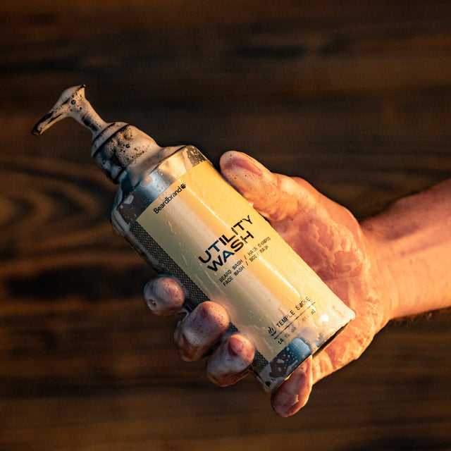 A person holding a sudsy container of Beardbrand Utility Wash in one hand, showing the front of the label, in warm lighting against a wooden background.