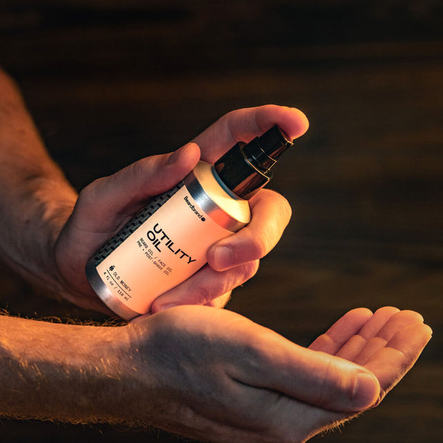 A person holding in their hand a bottle of Beardbrand Utility Oil and showing the front label. They are about to pump the product into their other hand. 