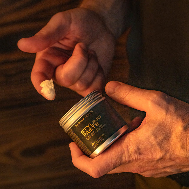 A person holding an open jar of Beardbrand Styling Paste showing the front label. They have scooped a small amount of Styling Paste out of the jar and you can see it on their finger. It is white and a marshmallowy texture.