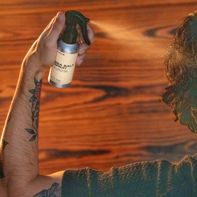 A person holding Beardbrand Sea Salt Spray, showing the front label, spraying it on their hair. The fine mist can be seen against a wooden background and warm lighting.