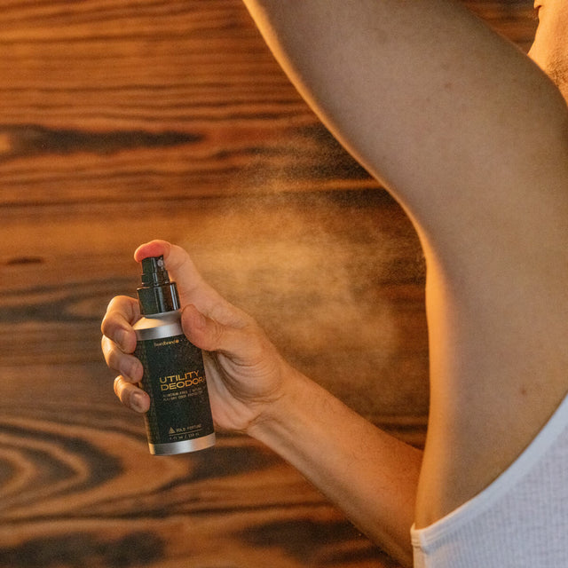 Man holds a bottle of Beardbrand Utility Deodorant with a black label and sprays it toward his underarm. The fine mist is captured mid-spray under warm lighting against a wooden background.