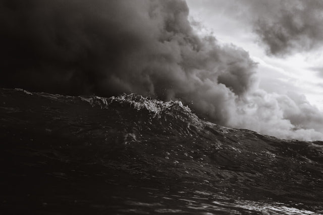 Black-and-white close-up of a dark ocean wave under ominous storm clouds, capturing the motion and texture of the turbulent water.