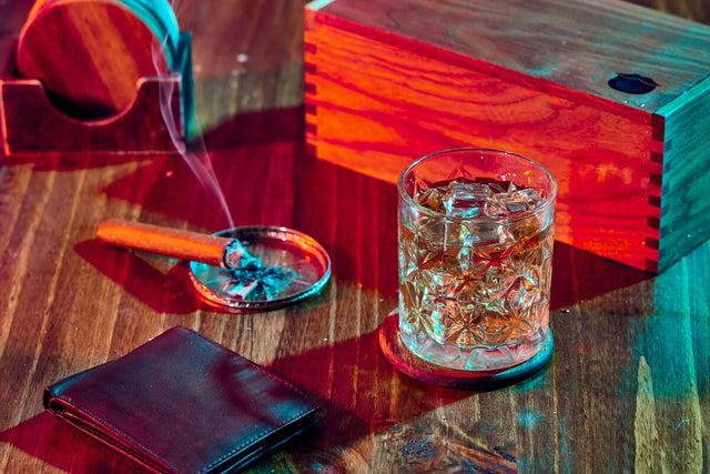 Glass of whiskey with ice on a wooden table, accompanied by a smoking cigar in an ashtray. A wooden box sits in the background, a leather wallet lies in the foreground.