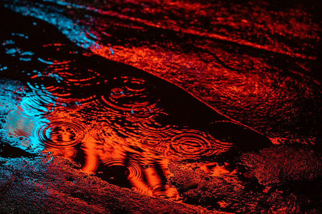 Puddle of water reflecting orange and blue neon lights, creating rippling concentric circles. Dark pavement with textured wet surfaces catching vibrant colors in a night scene.