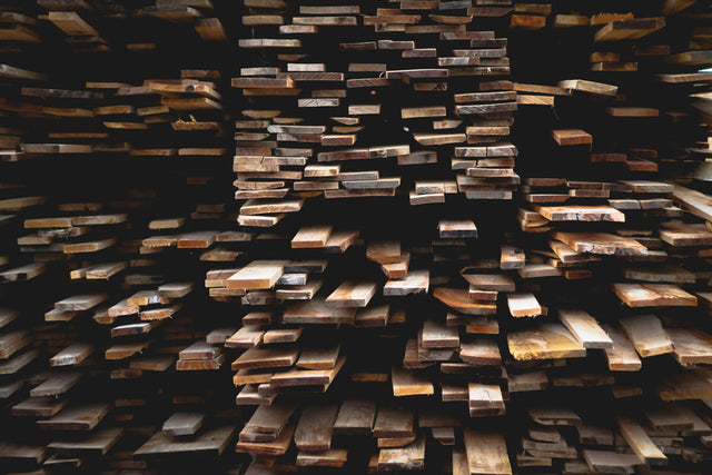 Stacked wooden planks arranged in rows inside a lumber yard or workshop. Various sizes of light brown timber boards are organized in layers, with natural wood grain visible in the warm lighting.