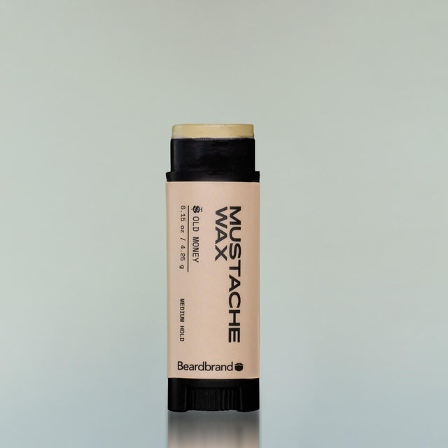 An Old Money Mustache Wax in a black plastic tube with the cap off showing the wax against a grey background.