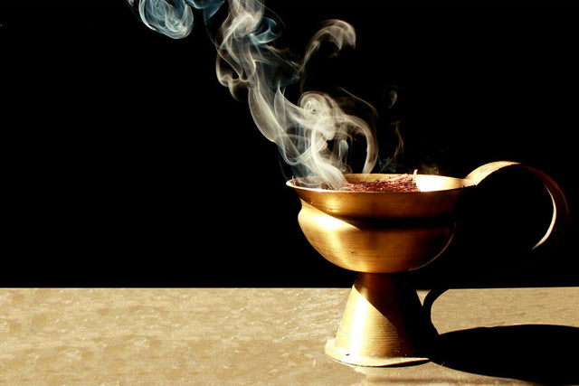 Golden brass incense burner with wisps of white and blue smoke rising against a black background. The ornamental vessel sits on a light beige surface, casting a soft shadow.