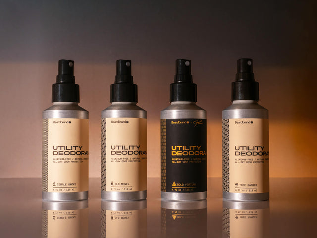 Four aluminum spray bottles of Beardbrand Utility Deodorant are displayed side by side on a reflective surface with a warm, gradient background.